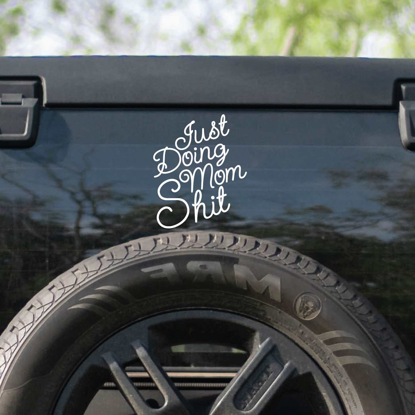 A white decal on the back of a tinted window of a Jeep that says, "Just doing mom shit.".